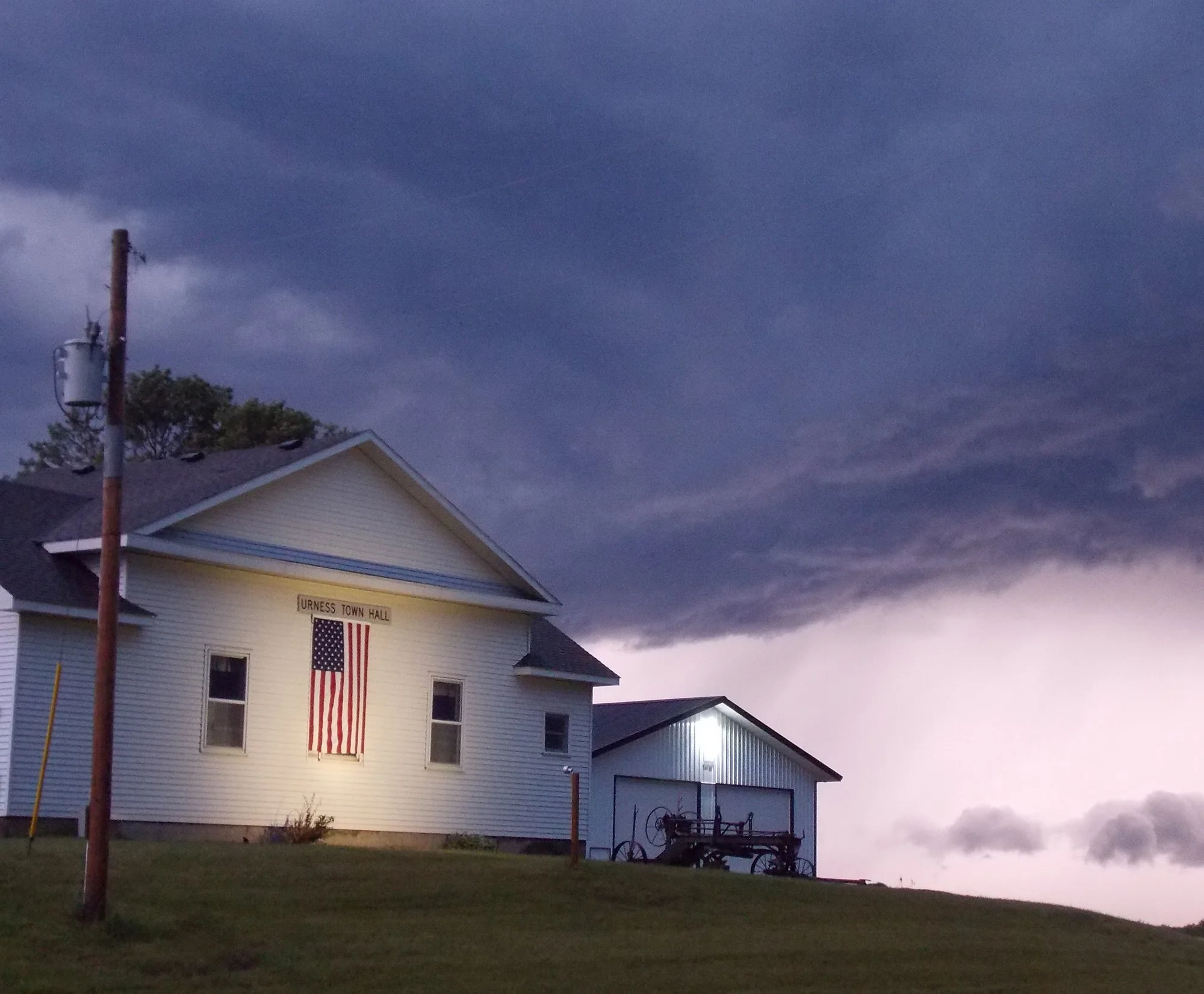 A white two-story house with an American flag hanging from a flagpole on the front. A sign on the house reads 'URNESS TOWN HALL.' There is a porch with outdoor furniture and a garden area in front. The sky is dark and cloudy, indicating evening or st