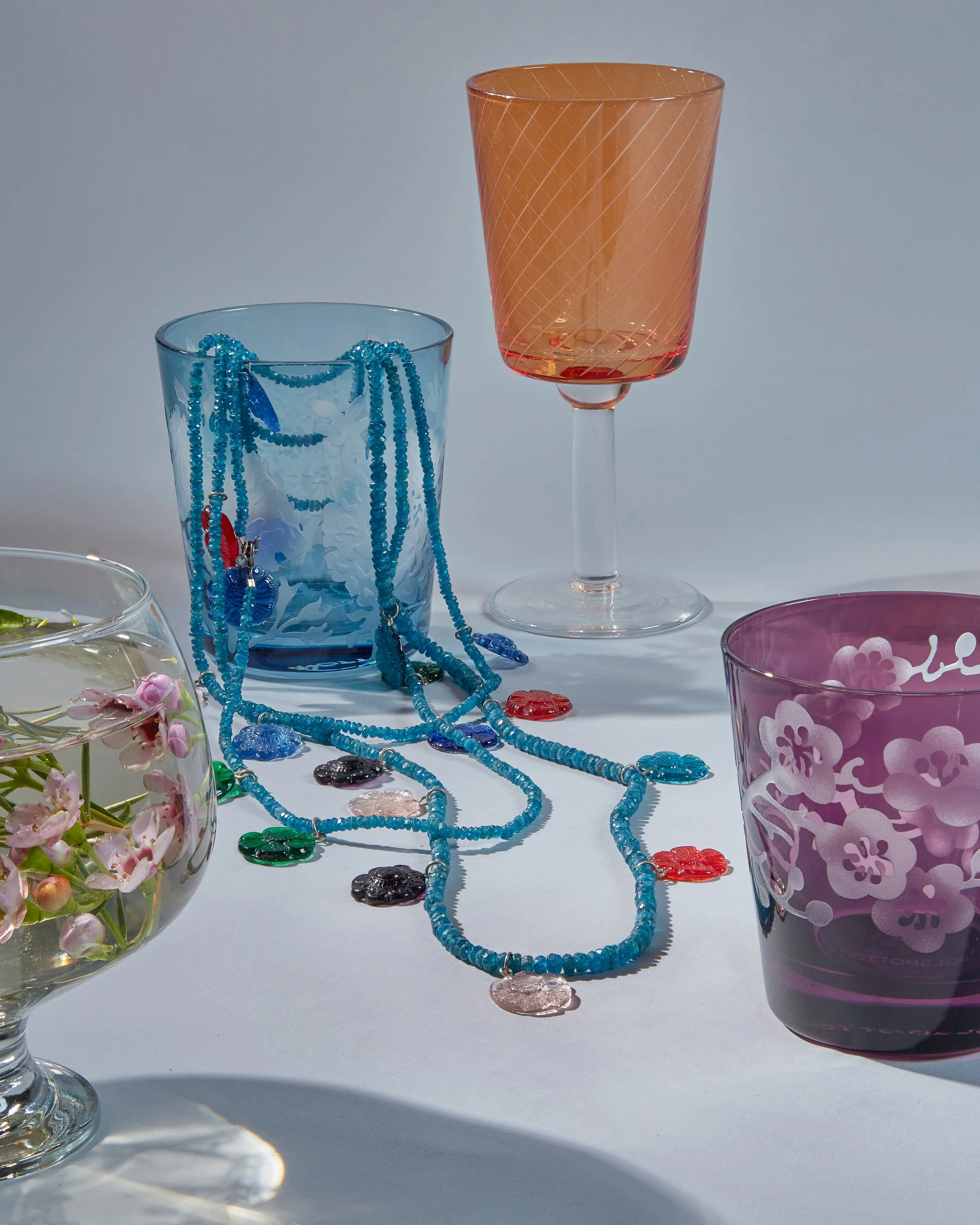A collection of glassware, including a tall smoky gray glass, clear glasses with floral etchings, a large bowl with a sunflower design, and assorted jewelry Polspotten X Ornamental Excess still life editorial advertising 
