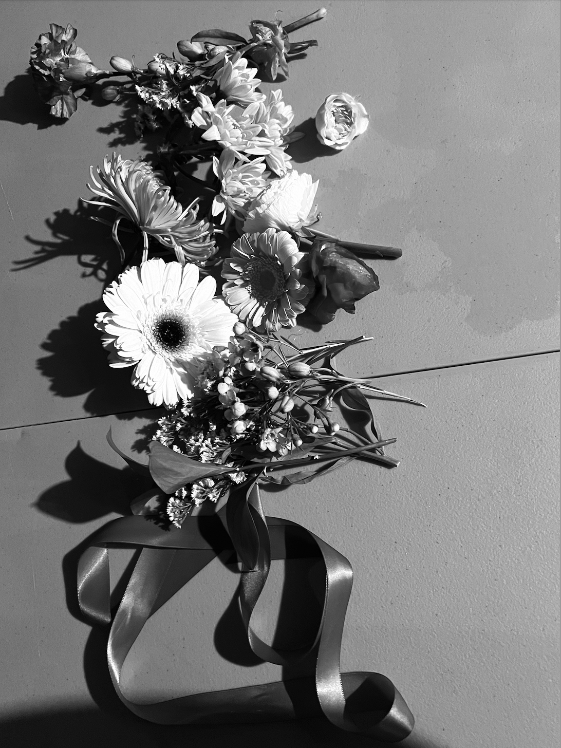 Black and white photograph of a bouquet of various flowers with a ribbon at the bottom, cast with shadows on a flat surface. Product Still life Photography by Gabrielle Gabe Weber and Hope Misterek, stylist