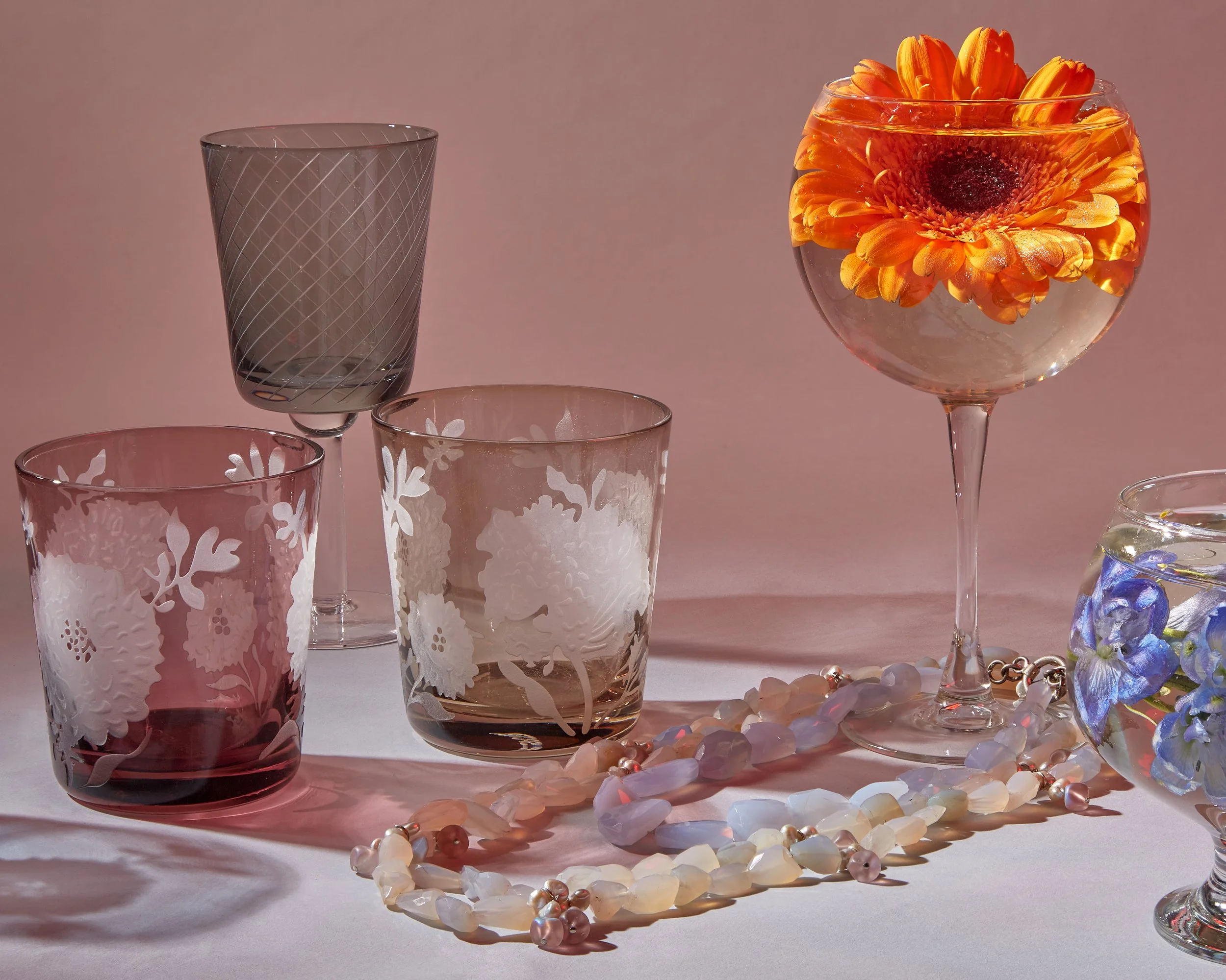 A collection of glassware, including a tall smoky gray glass, clear glasses with floral etchings, a large bowl with a sunflower design, and assorted jewelry Polspotten X Ornamental Excess still life editorial advertising 