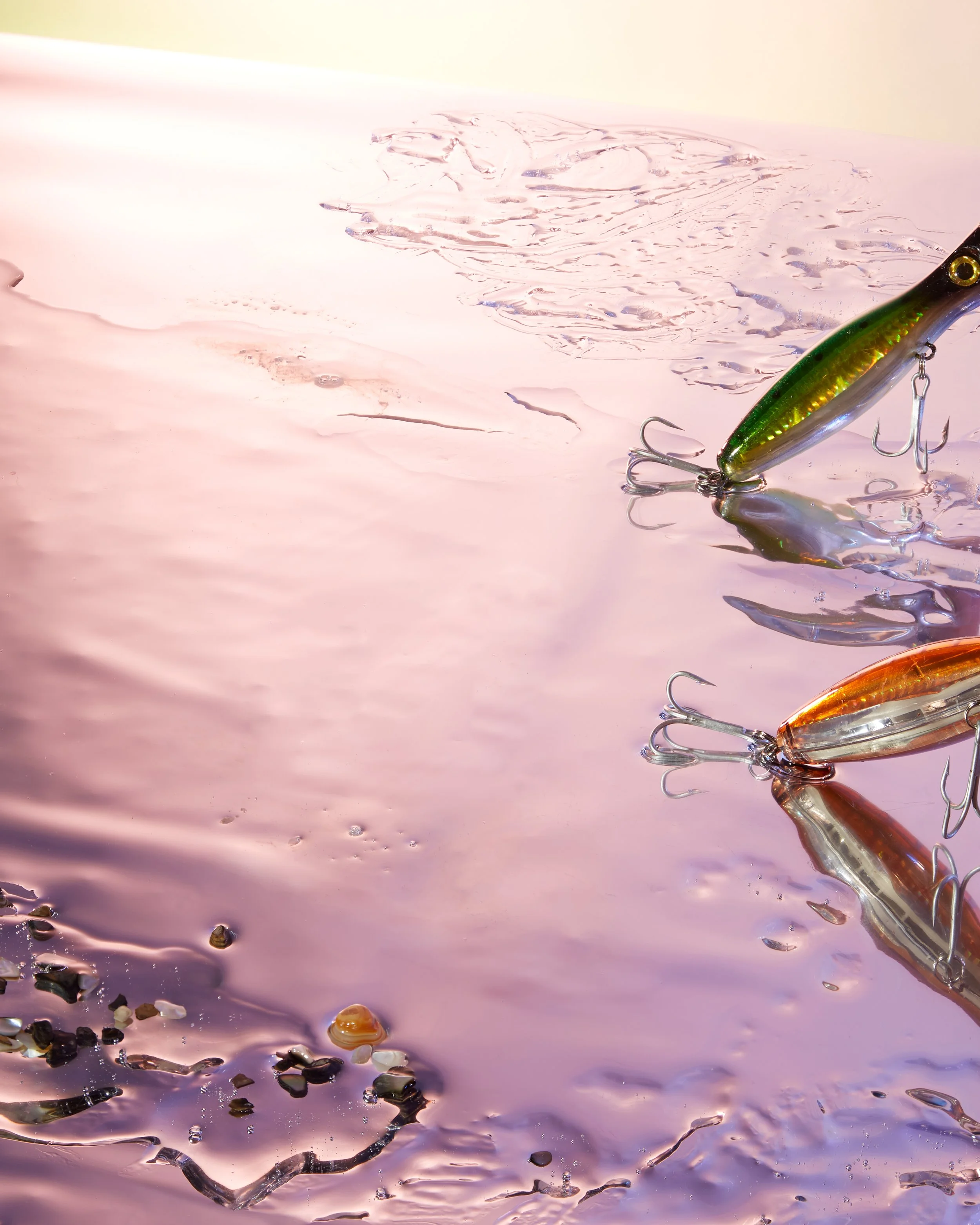 Close-up of a fishing lure in water with colorful reflections and scattered small shells or rocks along the surface. Creative Concepting Rapala and Stike King for advertising editorial, advertorial by Gabe Weber and Hope Misterek, branding, image, 