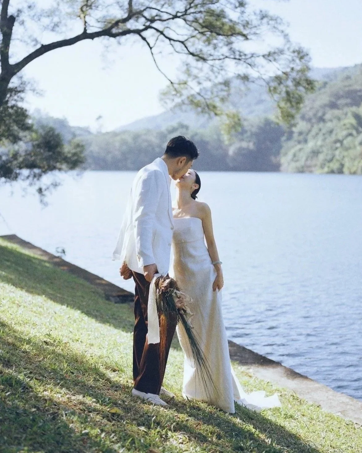 A couple dressed in wedding attire standing by a lakeside, sharing a kiss, with trees and mountains in the background.