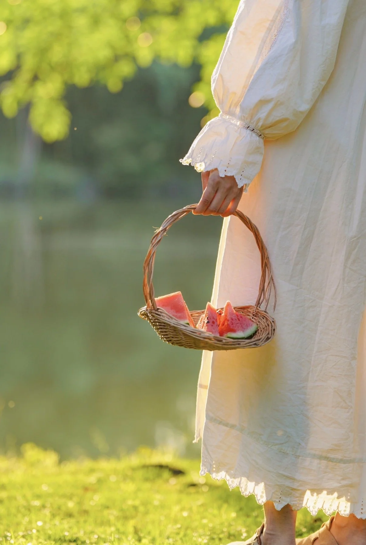 A person in a long white dress holding a small wicker basket with slices of watermelon, standing outdoors near a body of water with greenery in the background.