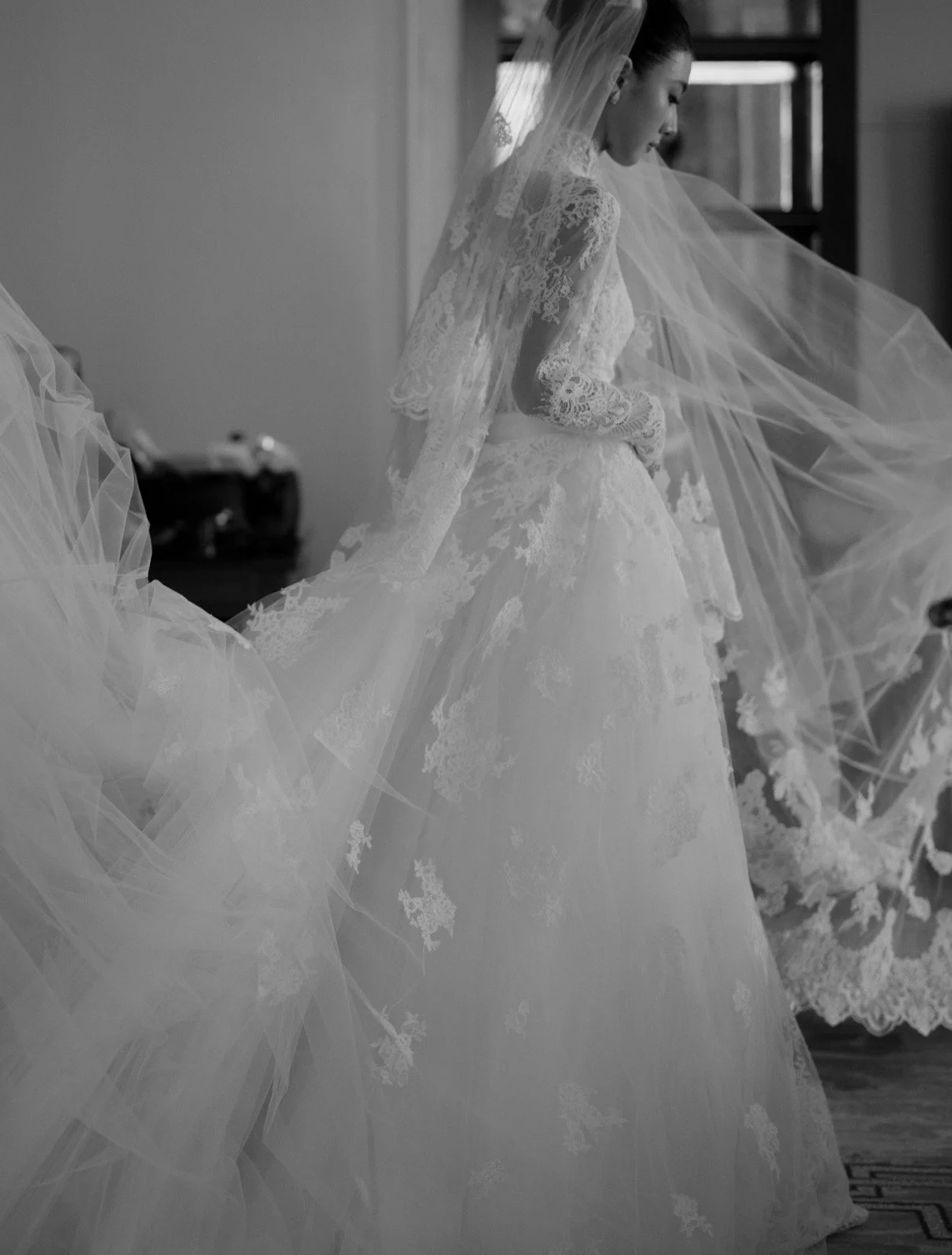 A woman in a wedding dress with lace details is adjusting her gown, with a long veil flowing around her.
