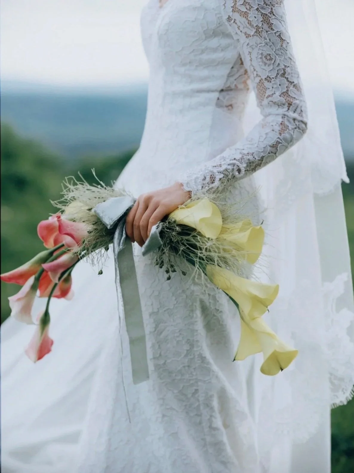A woman in a white lace dress holding a bouquet of pink, yellow, and peach-colored flowers, standing outdoors with a blurred green and blue background.