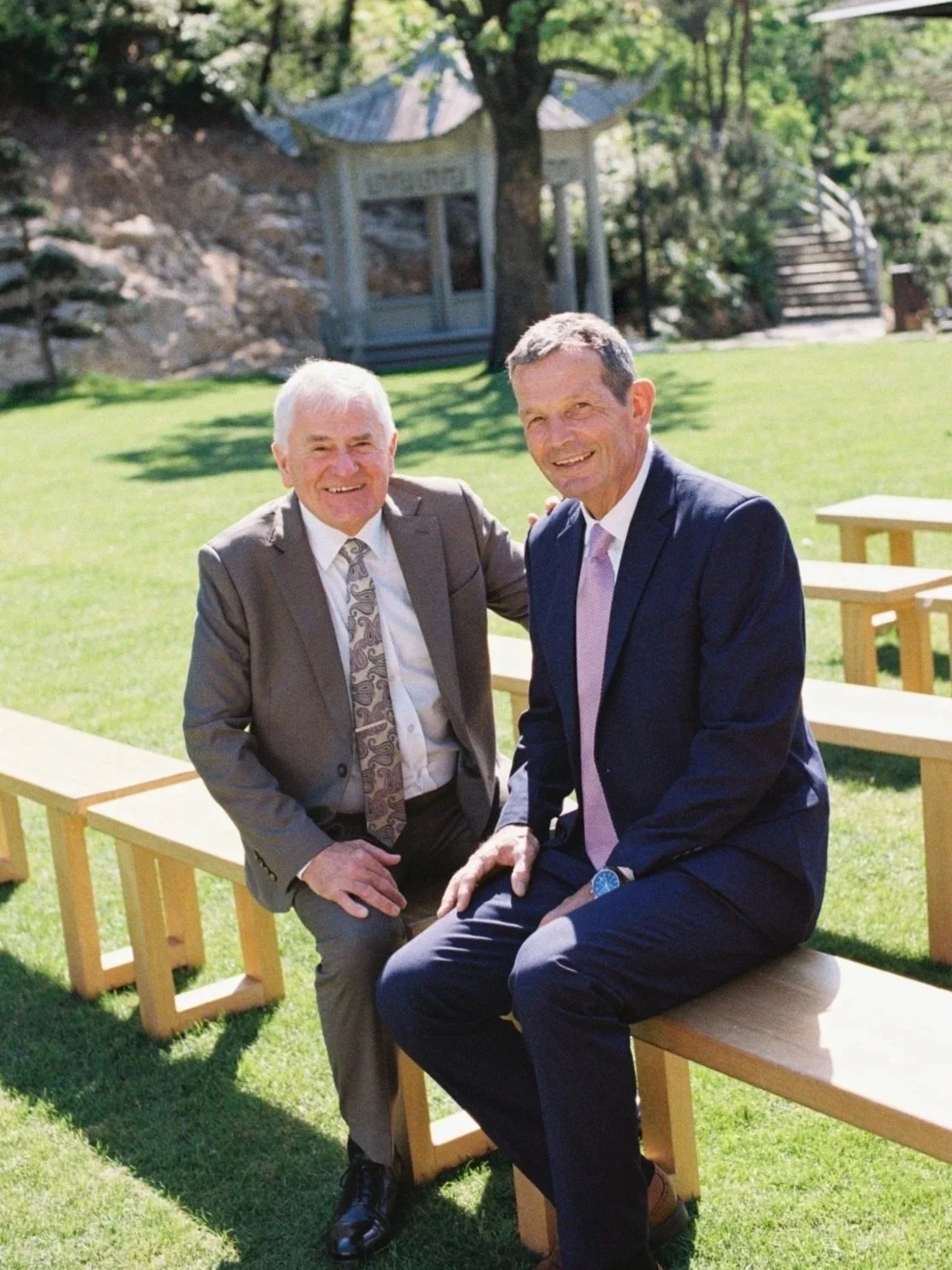 Two men in suits sitting on a wooden bench outdoors, smiling, with a grassy area, trees, and a small pavilion in the background.