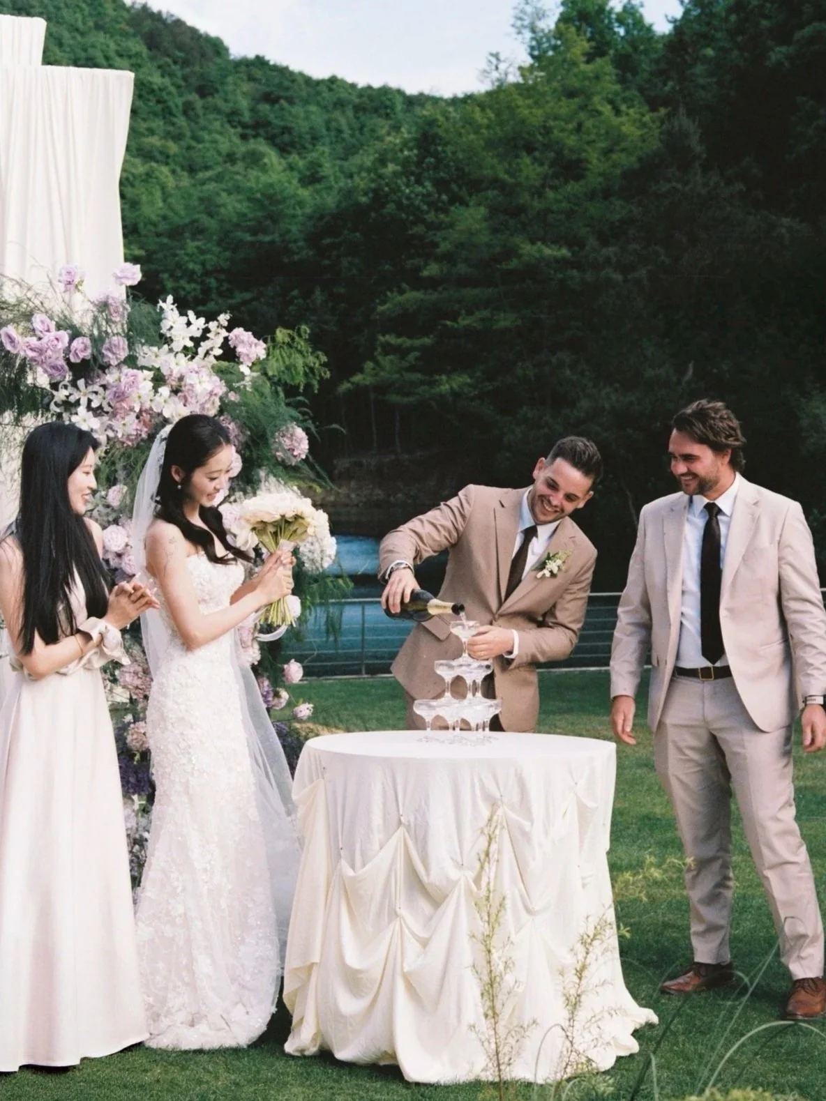 A wedding celebration outdoors with five people, two women and three men. The man in the center is pouring champagne into a tower of glasses, and the others are smiling. The scene features a scenic background of trees and a river, decorated with flow