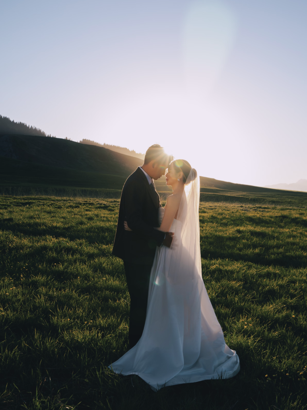 Couple in wedding attire standing closely together in a grassy field at sunset, with hills in the background.