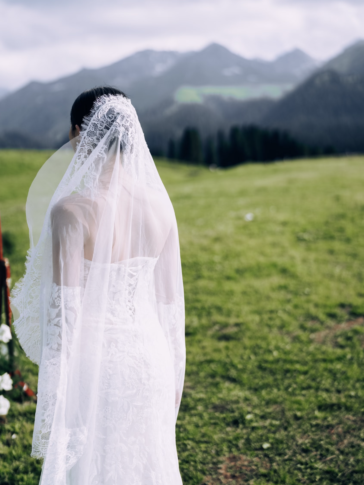 A woman in a white lace wedding dress with a lace veil standing outdoors on a grassy field with mountains in the background.