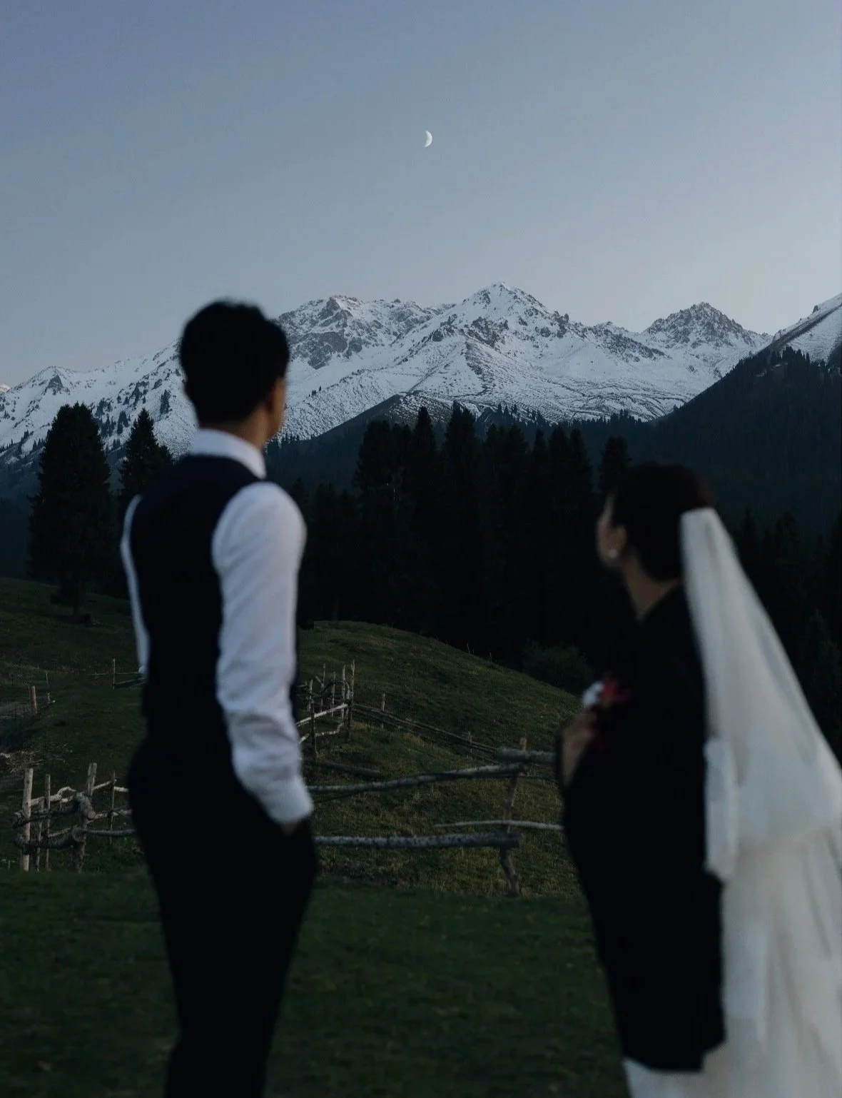 A bride and groom dressed in wedding attire standing outdoors, facing snowy mountains and a crescent moon in the evening sky.