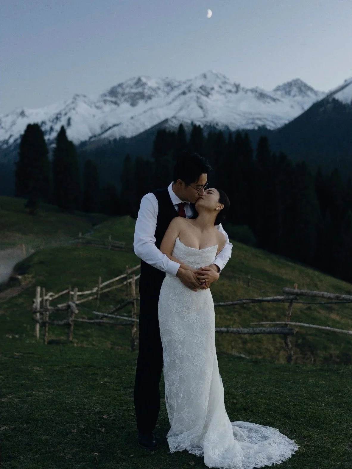 A couple, dressed in wedding attire, sharing a kiss outdoors during twilight with snow-capped mountains and a crescent moon in the background.