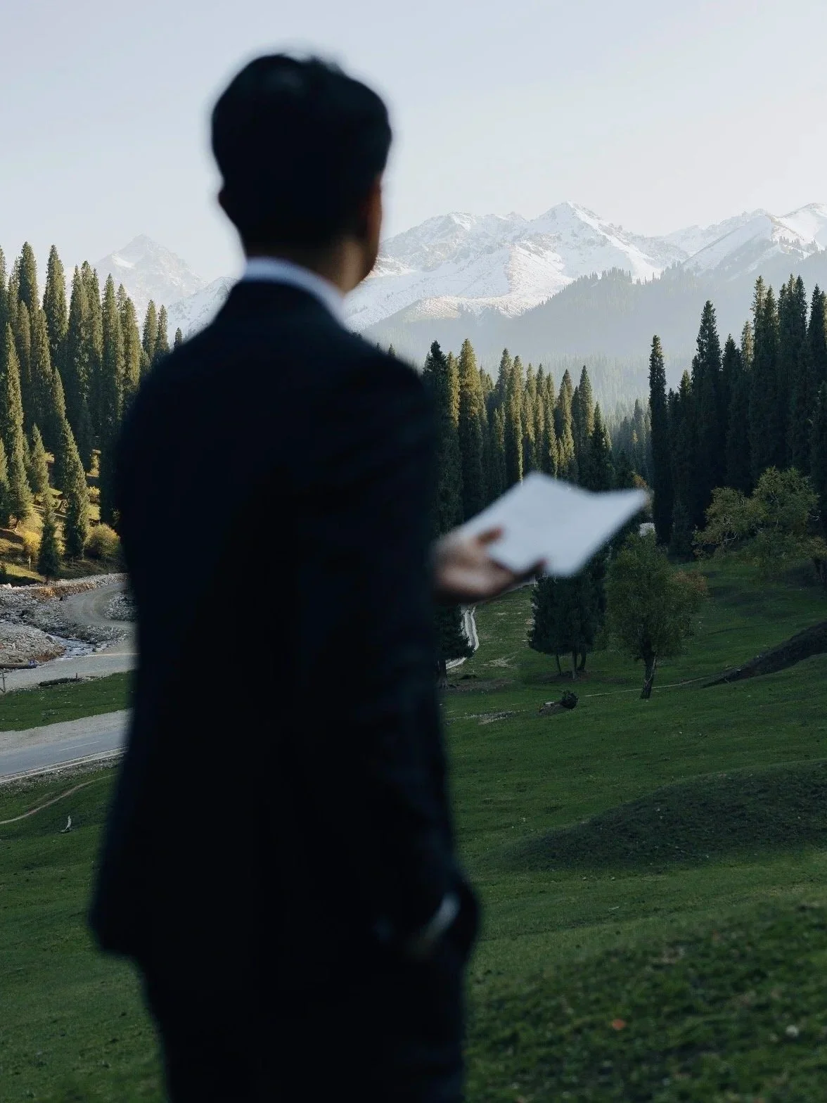 A man in a black suit holding a tablet, standing outdoors in front of a landscape of pine trees and snow-capped mountains.