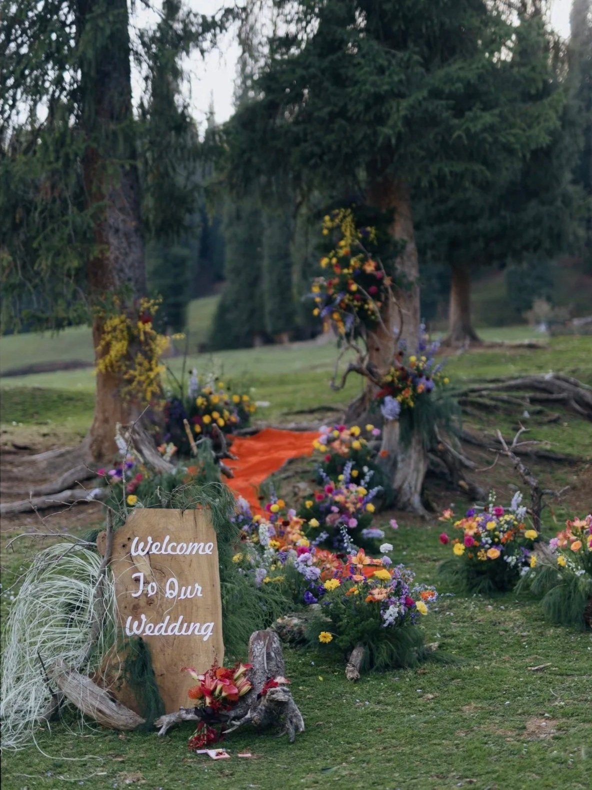 Outdoor wedding setup with colorful flowers arranged around tree trunks and branches, with a wooden sign reading 'Welcome To Our Wedding' at the front.