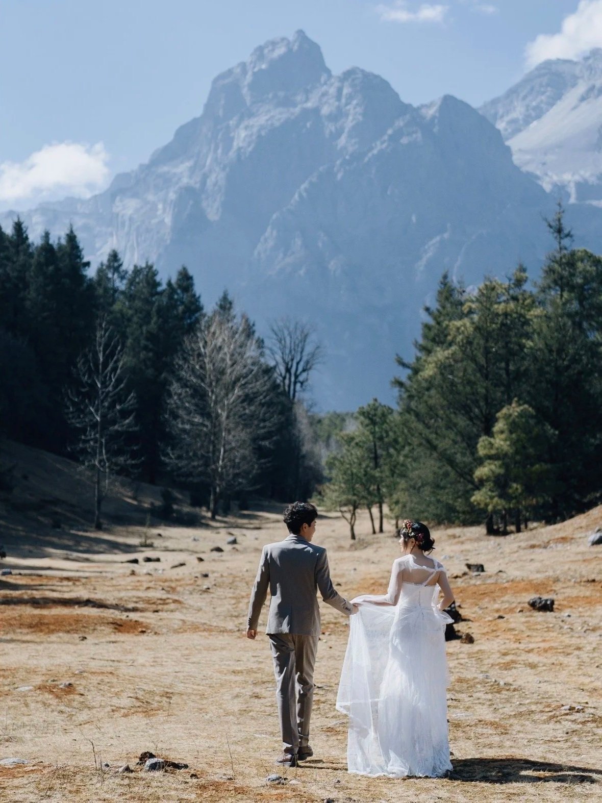 A bride and groom holding hands in a mountain landscape with trees, mountains, and a clear sky in the background.