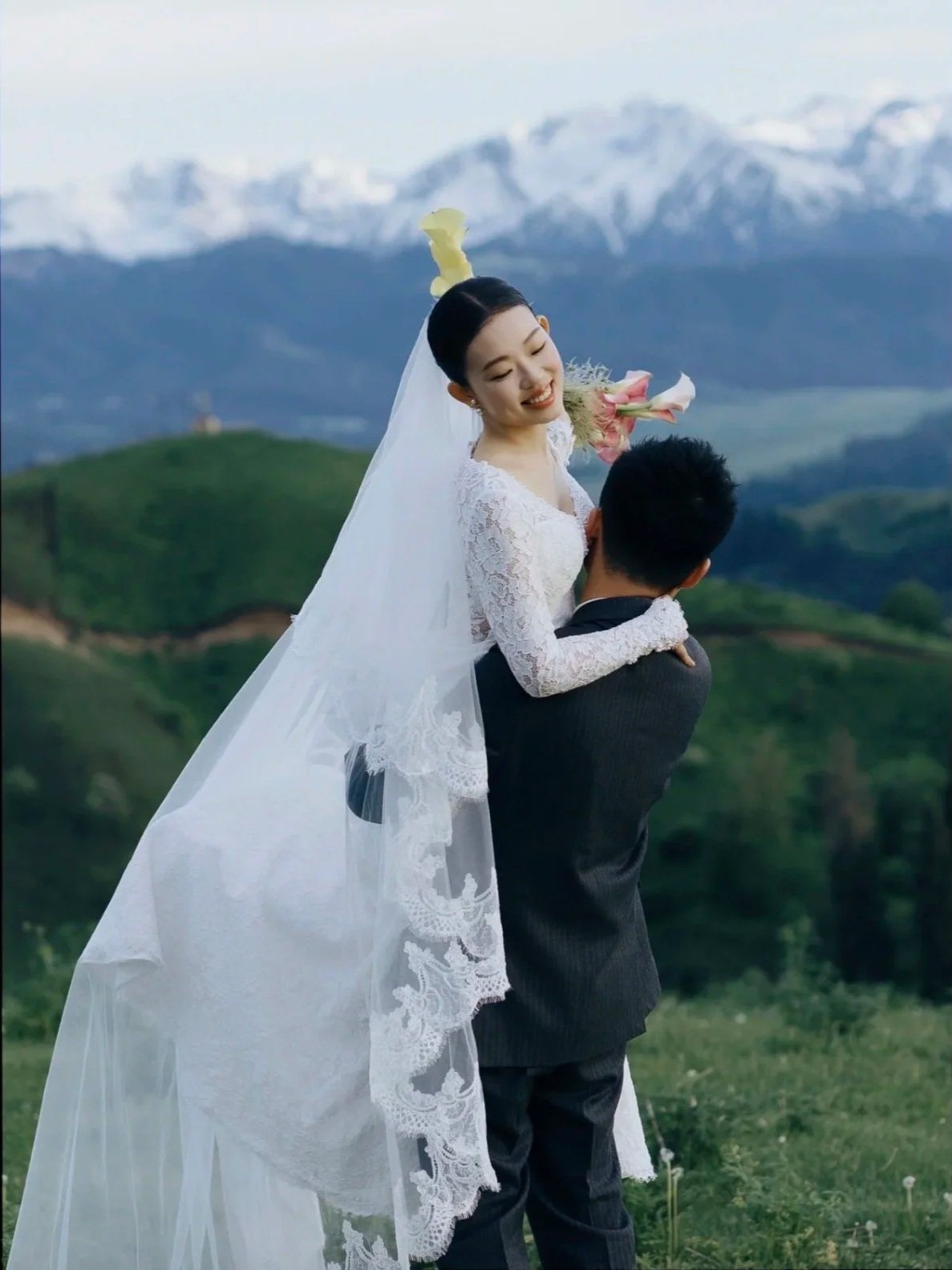 A bride and groom outdoors in a mountainous landscape with snow-capped peaks, the bride smiling and holding a bouquet, being lifted by the groom.