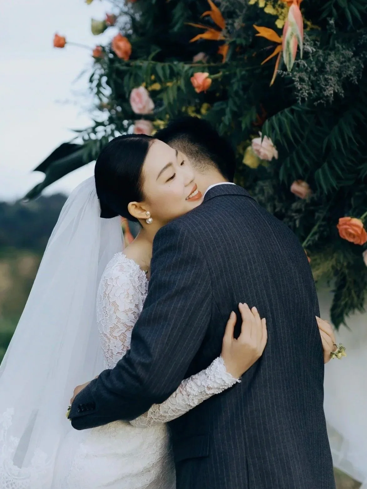 A bride and groom embrace outdoors with flowers and greenery in the background.