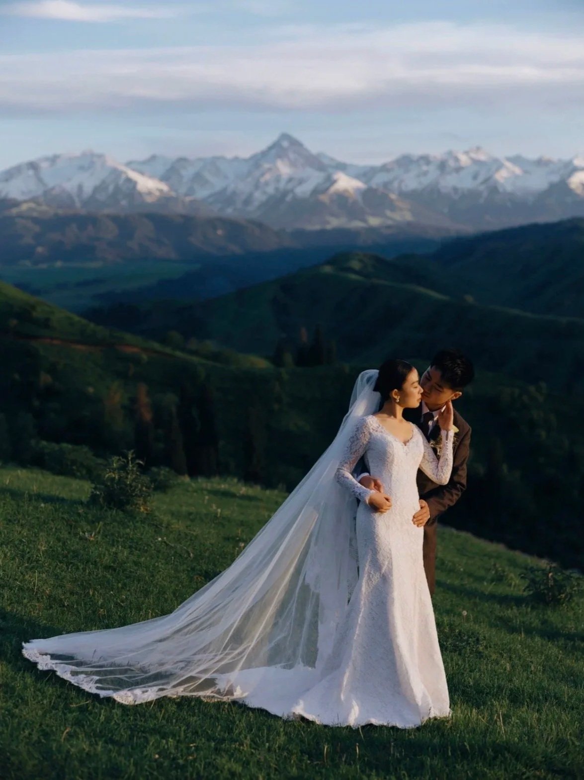 A bride and groom sharing a romantic moment outdoors on green grass with a mountain range with snow-capped peaks in the background.
