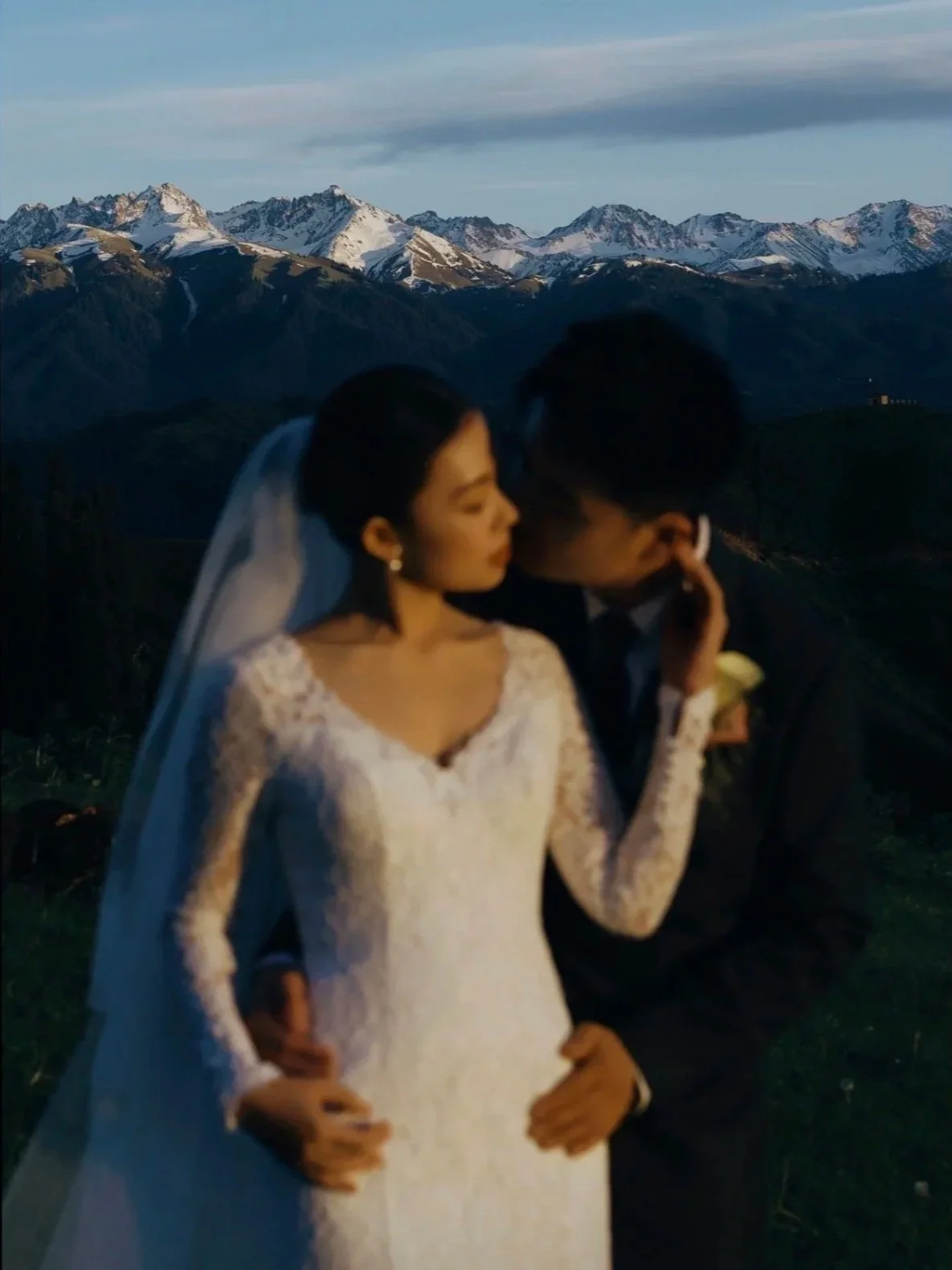 A bride and groom sharing a kiss outdoors with snow-capped mountains in the background at sunset.