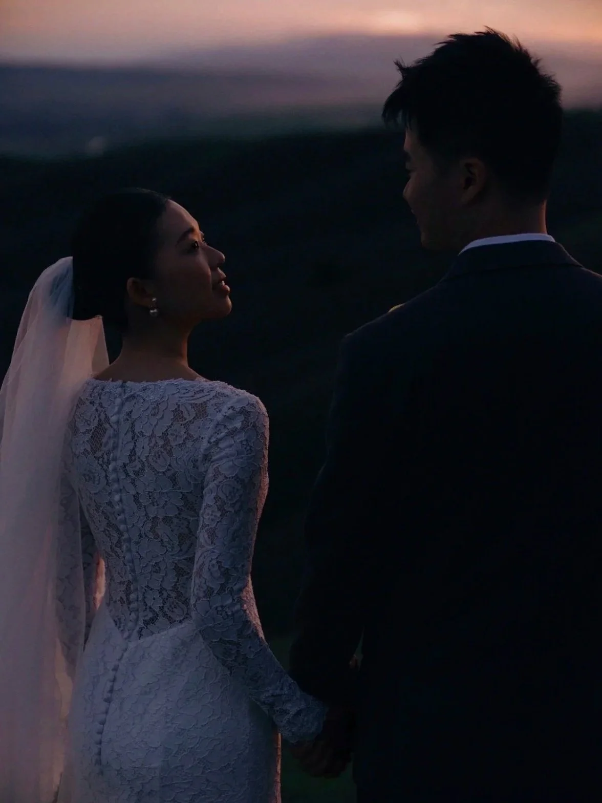 A bride and groom holding hands outdoors at sunset, looking at each other.