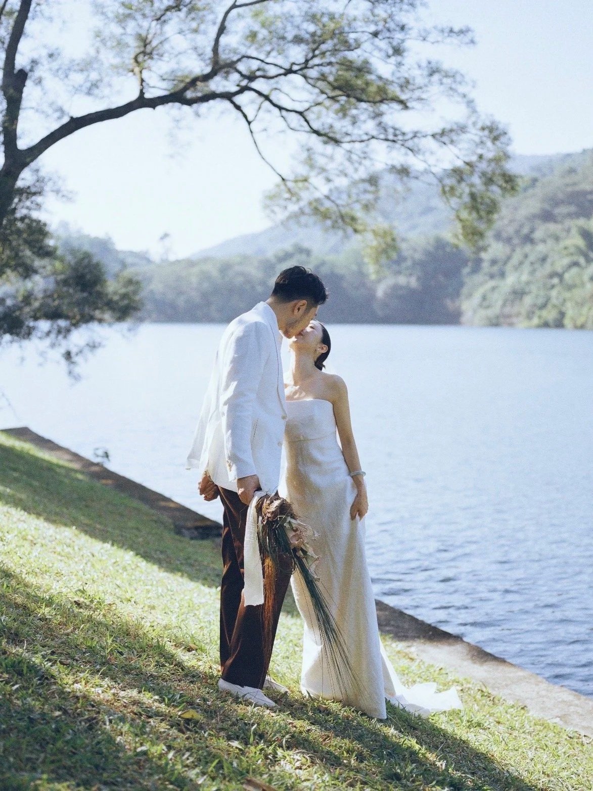 A couple dressed in wedding attire sharing a kiss by the water's edge with trees and mountains in the background.