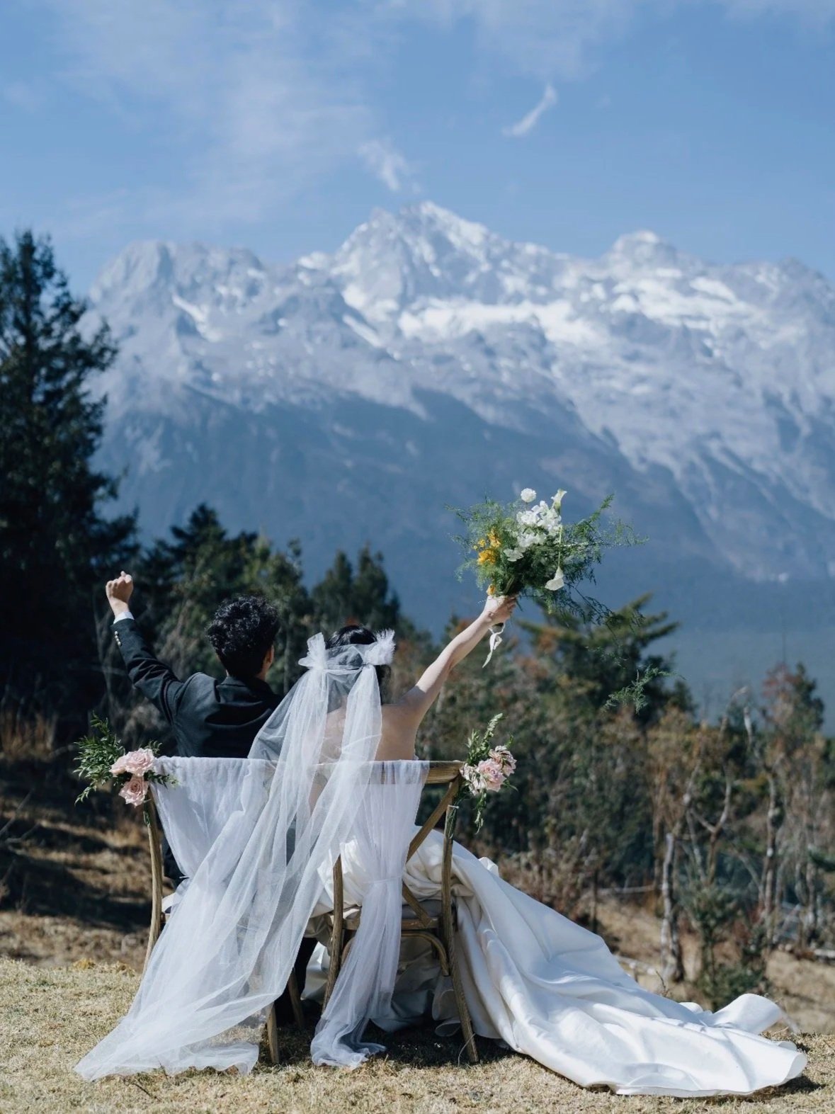 A bride and groom sitting outdoors on a wooden bench, with the bride holding a bouquet and raising it in celebration, against a backdrop of snow-capped mountains and trees.