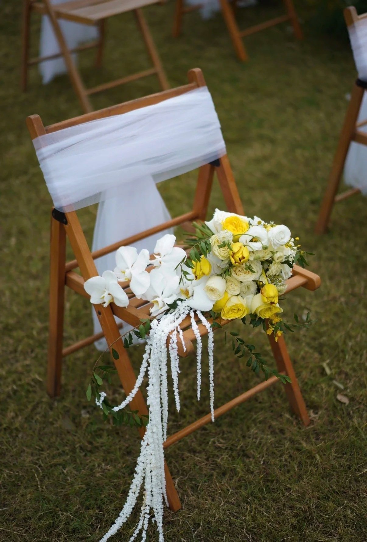 A wooden chair decorated with white fabric and a floral arrangement of white and yellow flowers with greenery, set outdoors on grass for a wedding or special event.