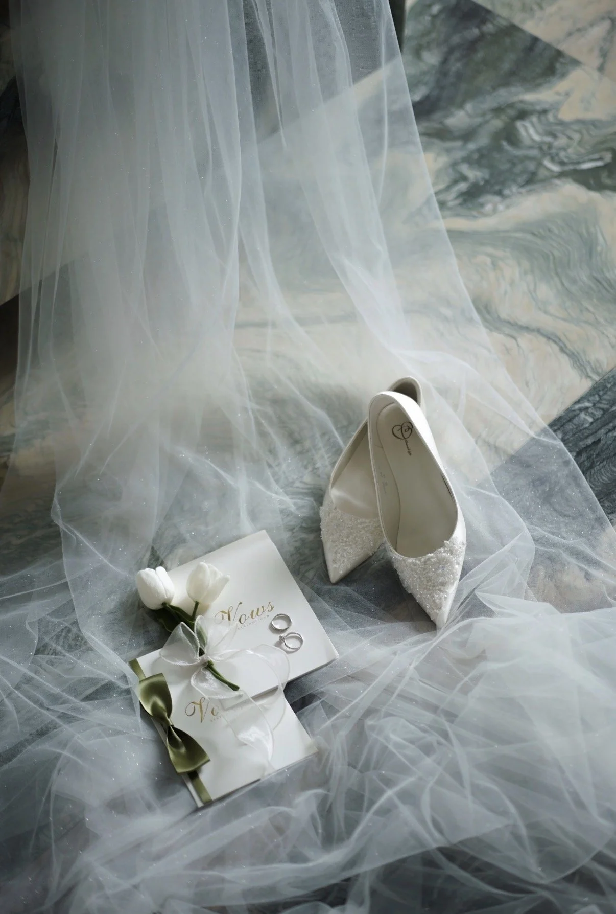 Wedding accessories including white high heels with lace embellishments, a white bouquet with flowers, a ring, and a greeting card with the word 'Nous' surrounded by sheer white fabric on a marble surface.