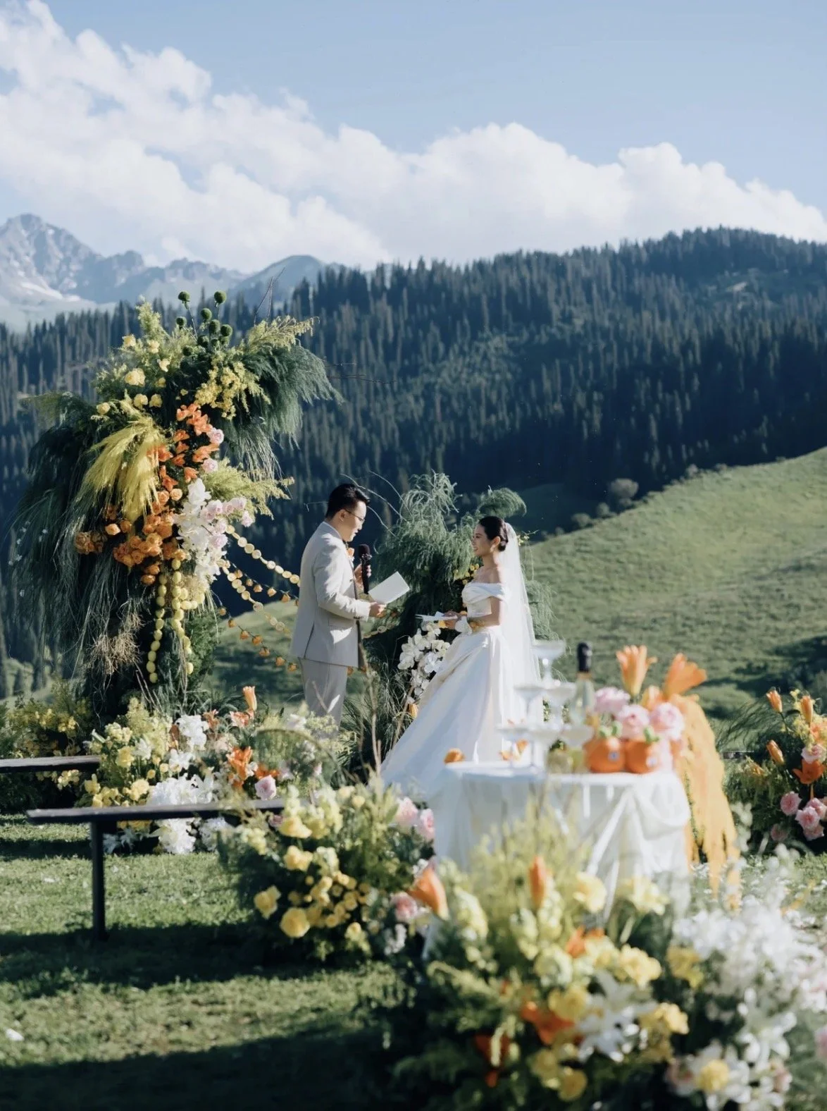 A wedding ceremony taking place outdoors with mountains and forest in the background. A bride and groom stand in front of a floral arch, exchanging vows. The scene is decorated with colorful flowers and greenery, with a table set nearby.