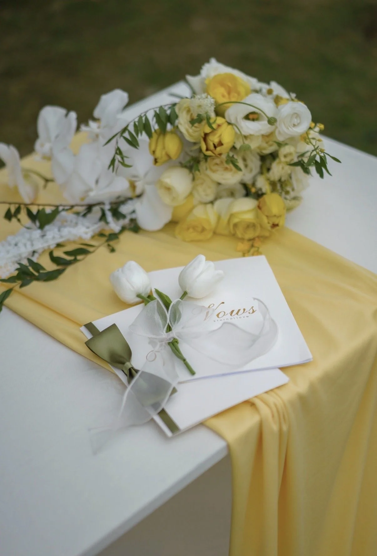 A wedding bouquet with white and yellow flowers and greenery, resting on a yellow fabric on a table. A white card with gold writing and a small white flower bouquet tied with a white ribbon are placed nearby.
