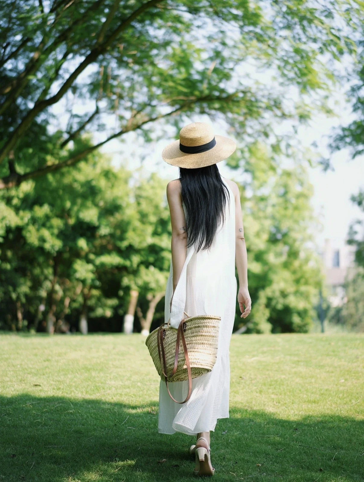 A woman wearing a white dress, a wide-brimmed hat, and sandals, carrying a woven handbag, walking in a lush green park with trees and grass.