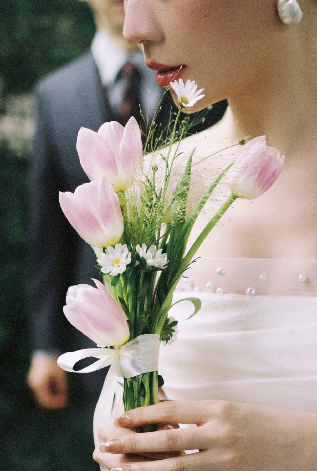 A bride holding a bouquet of pink tulips, white daisies, and greenery at her wedding, with a woman in the background wearing a pearl earring and wedding dress.