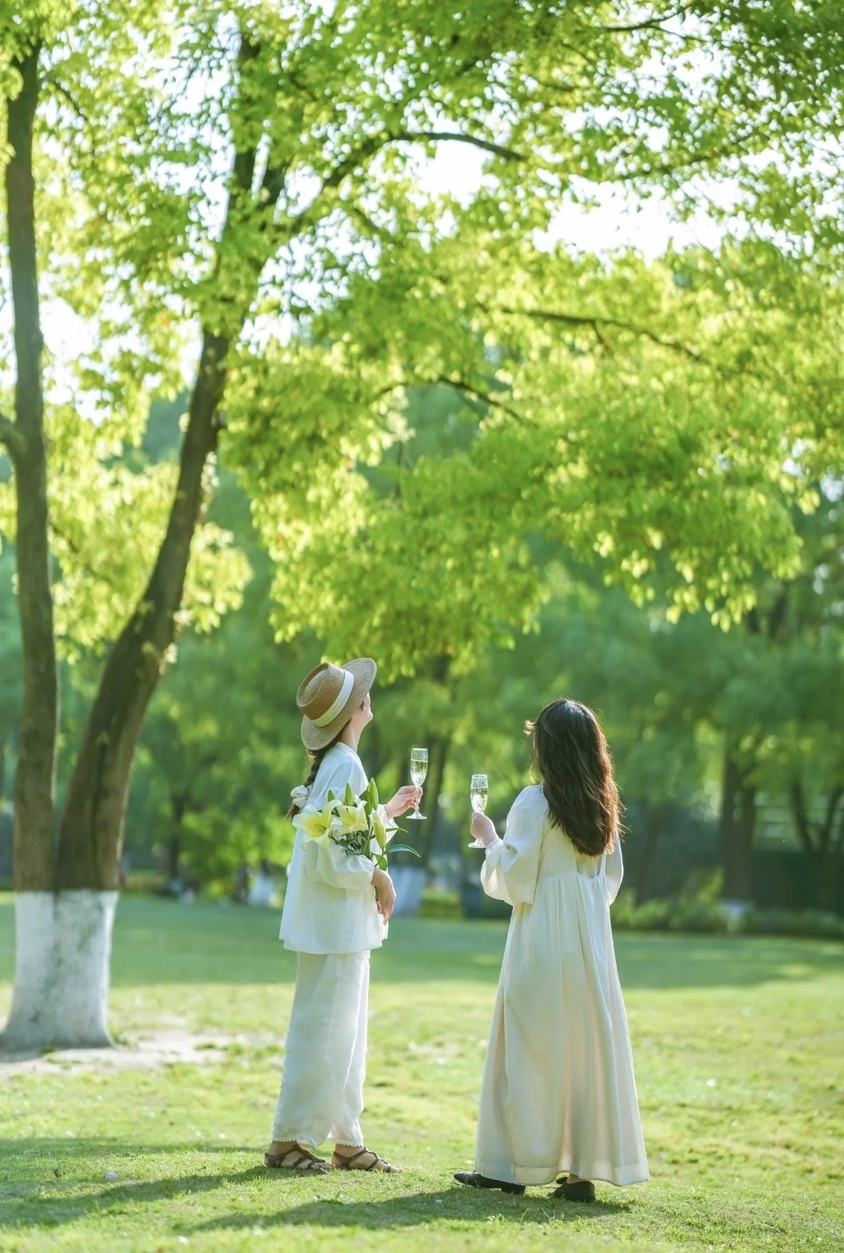 Two women in white dresses standing in a park with green trees, holding glasses of champagne and engaging in conversation. One woman also has a bouquet of white lilies.