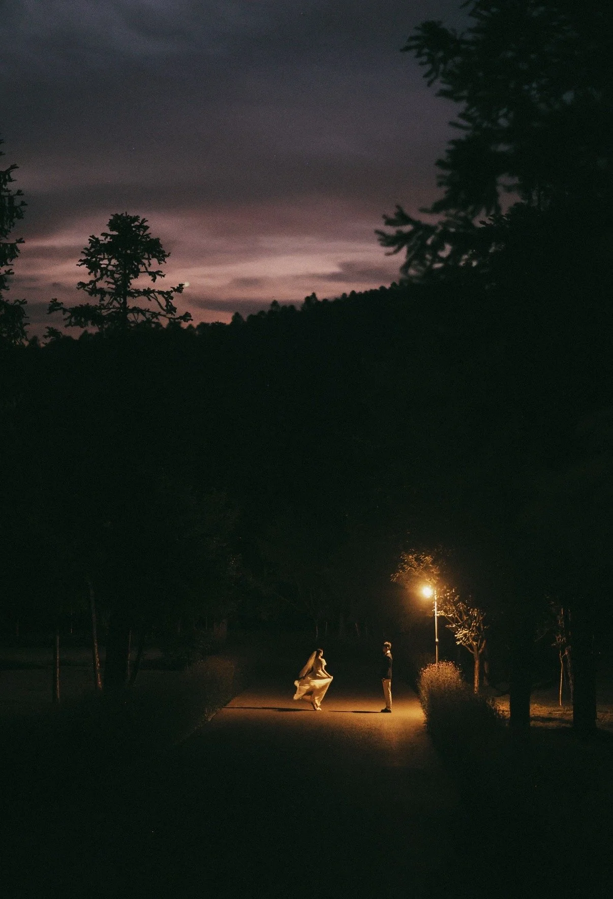A dark outdoor scene at night with a cloudy sky and trees. In the foreground, two people are standing under a streetlamp, with one person wearing a white dress that appears to be floating or moving.