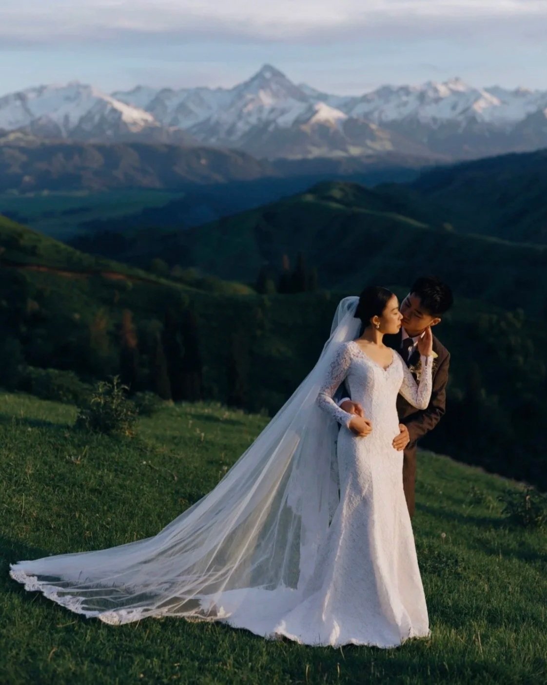 A bride and groom sharing an intimate moment outdoors with a picturesque mountain landscape in the background. The bride is wearing a white lace wedding dress with a long veil, and the groom is dressed in a dark suit.
