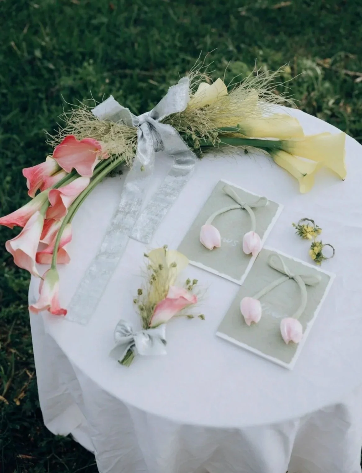 A white tablecloth-covered table with a floral arrangement including pink and yellow flowers, tied with a silver ribbon. There are two sets of ring boxes with wedding rings, pink flower petals, and small flowers arranged on the table. The background is grass.