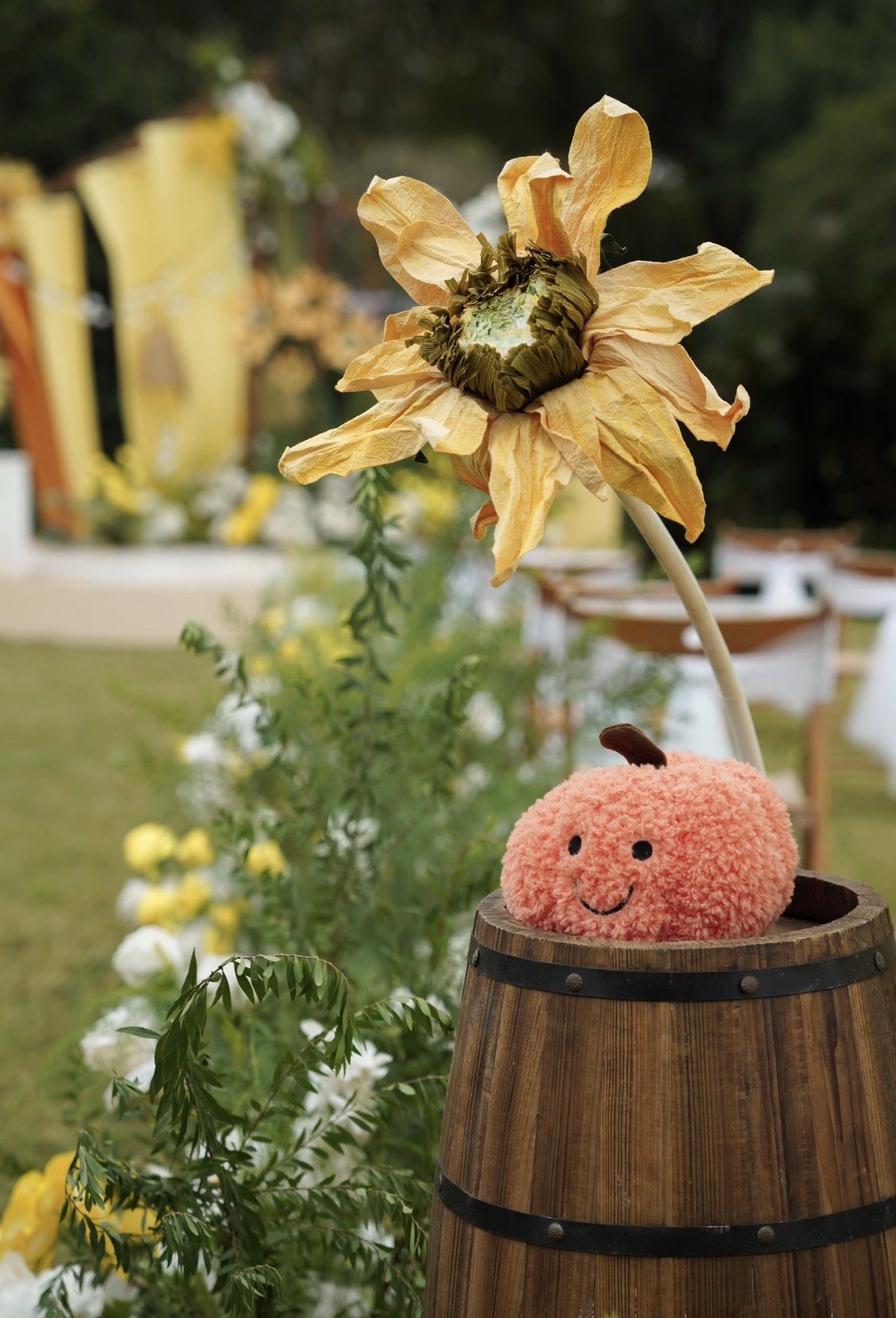 A pink, fuzzy stuffed toy with a smiling face and a small stem on top, sitting inside a wooden barrel, with a large dried flower with yellow petals behind it, set in an outdoor garden.