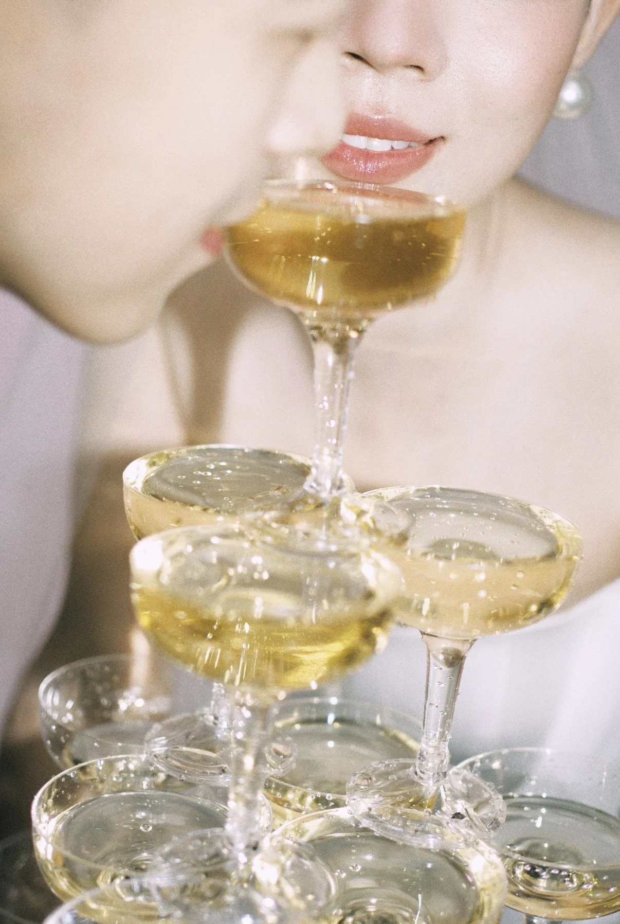 Close-up of a woman with glossy lips and pearl earrings, sipping champagne from a glass with multiple champagne glasses stacked in a pyramid formation.