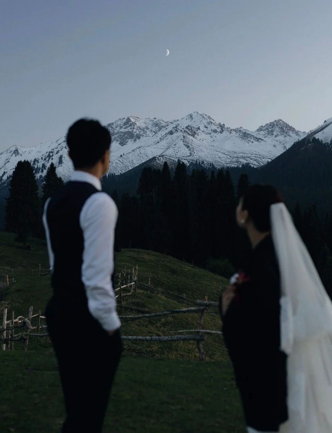 A couple, dressed in wedding attire, stands outdoors at dusk with snow-covered mountains and a crescent moon in the background.