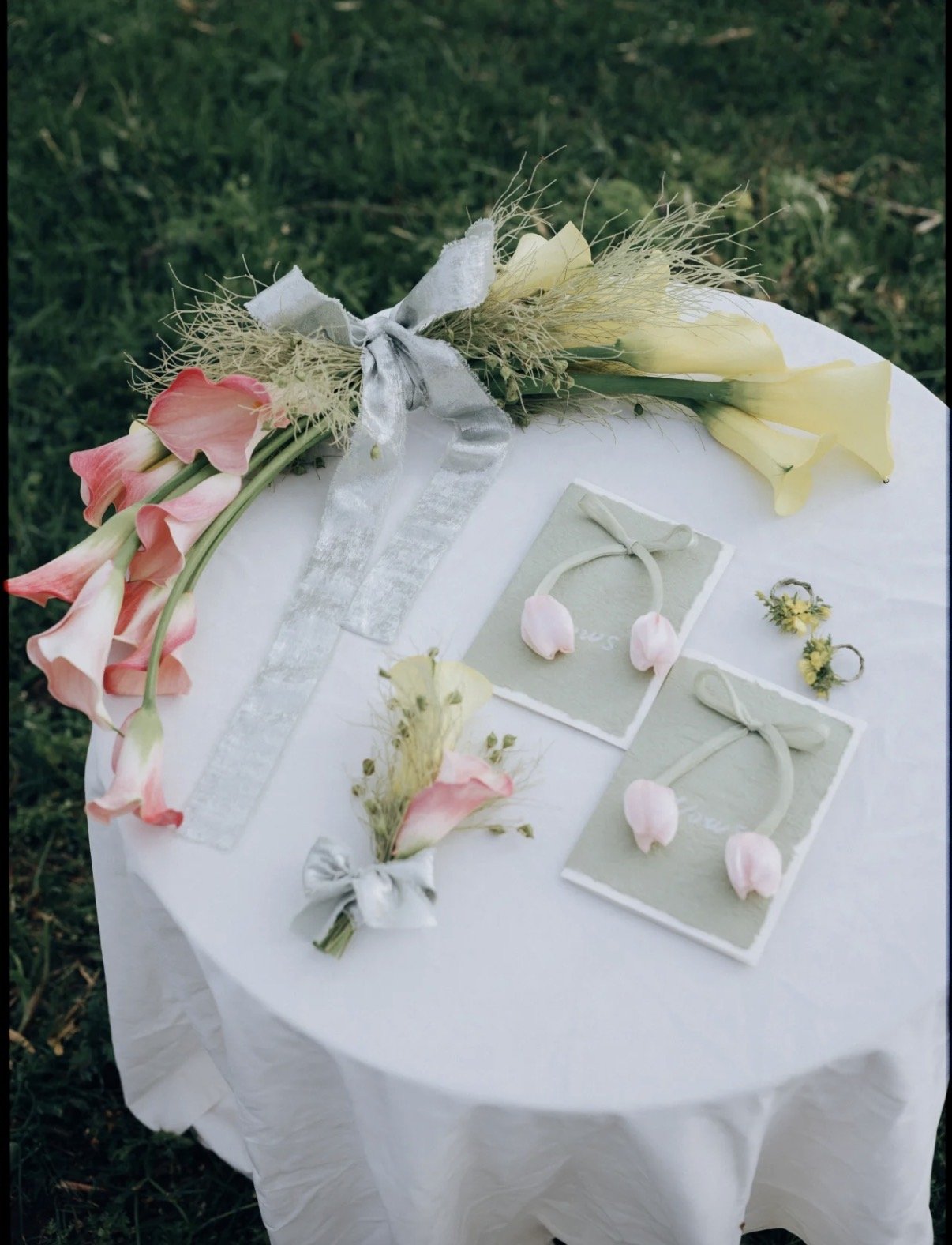 Table decorated with pink and yellow flowers, small jewelry pieces with flower accents, and white ribbons, set outdoors on grass.