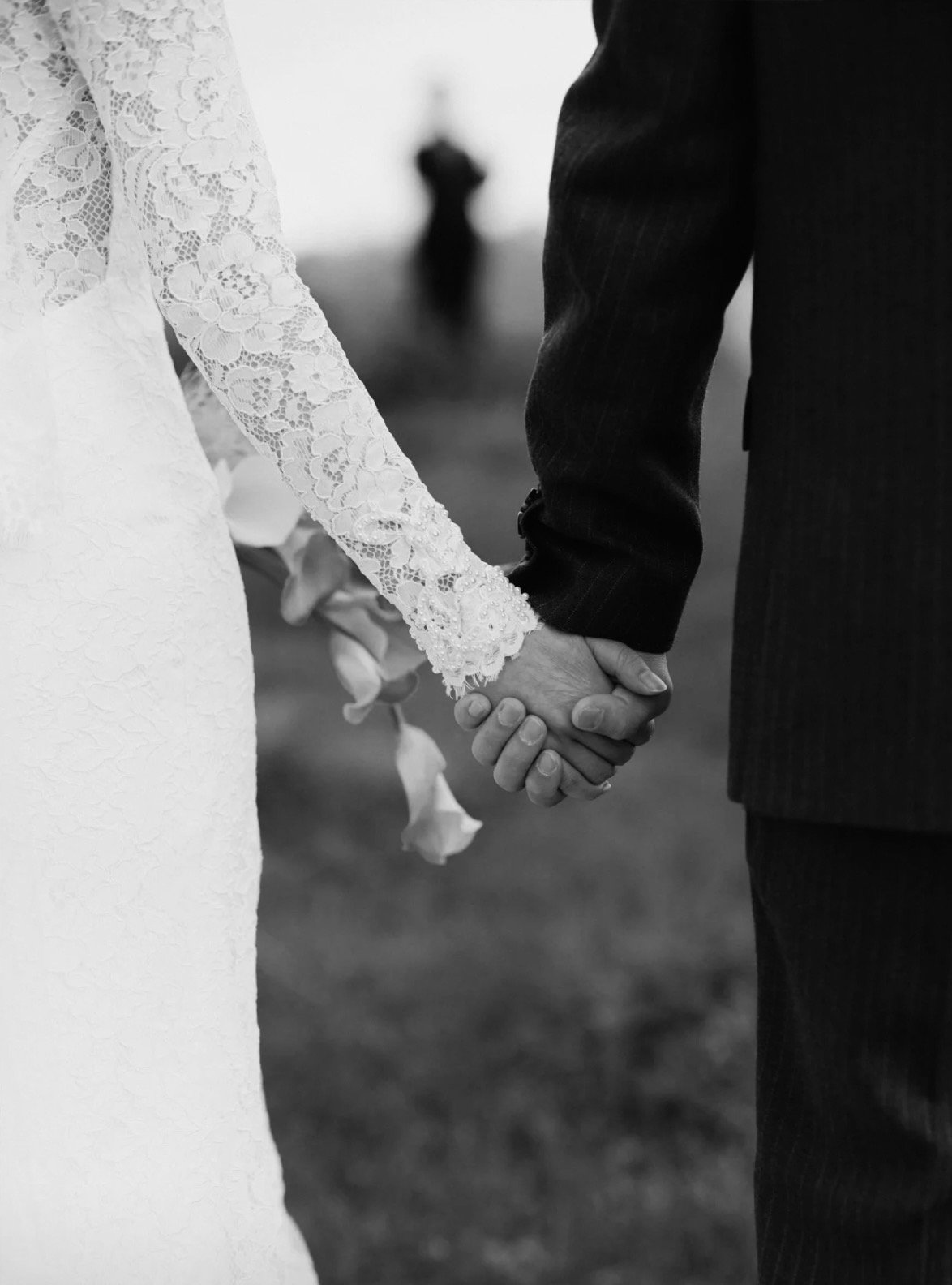 Close-up of a bride and groom holding hands during their wedding, with the bride wearing a lace dress and holding a bouquet, in black and white.