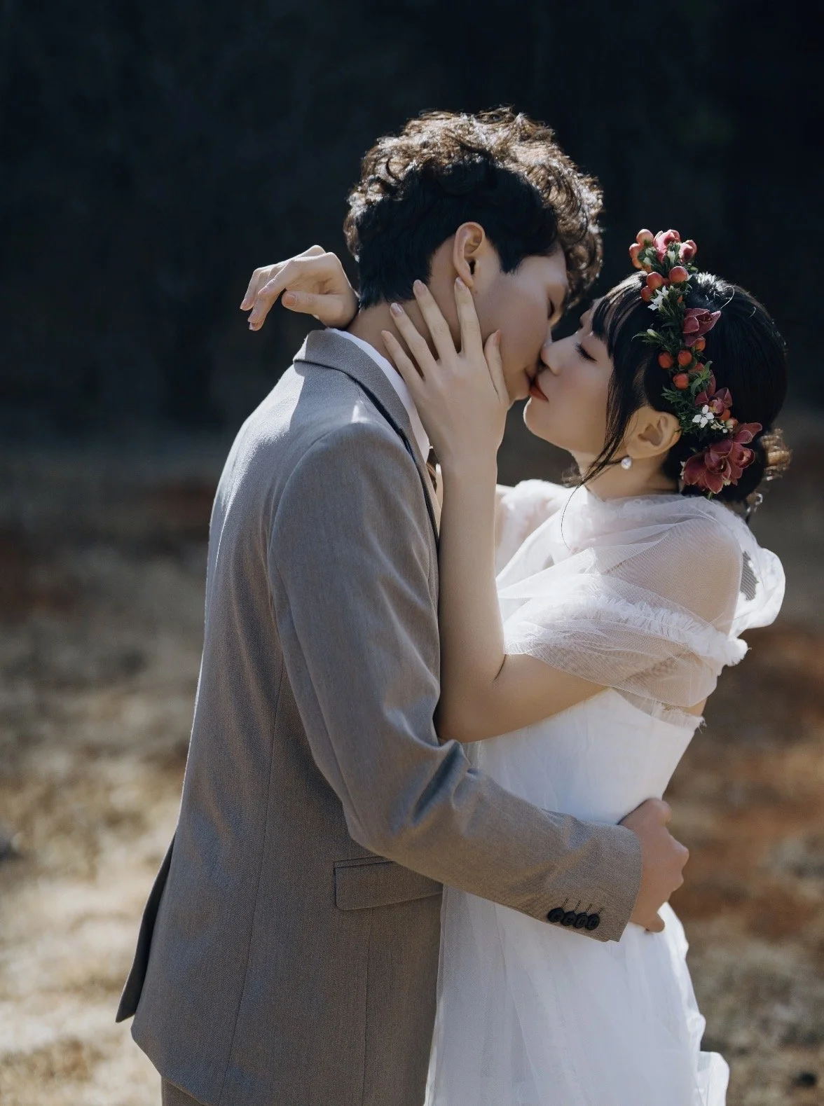 A couple in wedding attire sharing a kiss outdoors. The woman is wearing a white dress with puffy sleeves, a floral headpiece, and earrings. The man is in a gray suit. The woman has her hand on the man's face, and they are embracing each other.