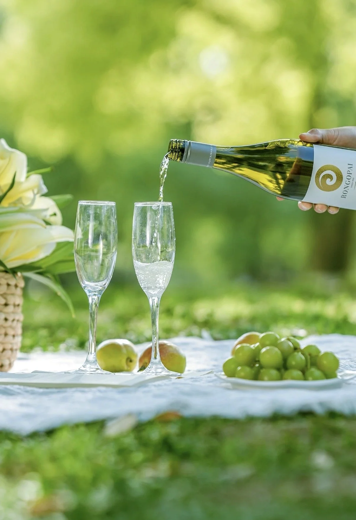 A hand pours white wine into a champagne flute on a picnic table with a backdrop of green trees, a bunch of green grapes, apples, and a flower arrangement in a basket.