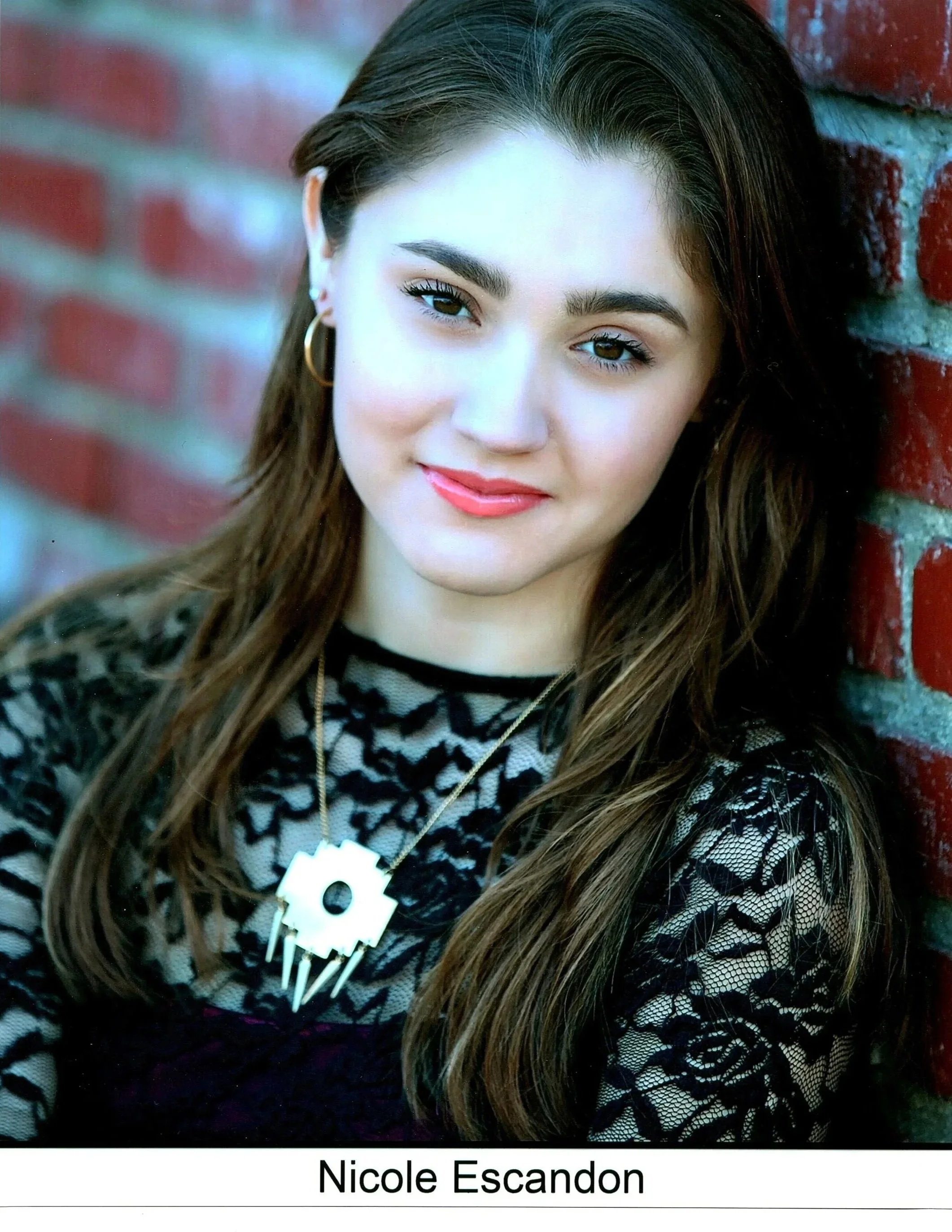 A close-up portrait of Nicole Escadon, a young woman with long brown hair, wearing gold hoop earrings and a black top with a lace pattern, standing against a red brick wall.