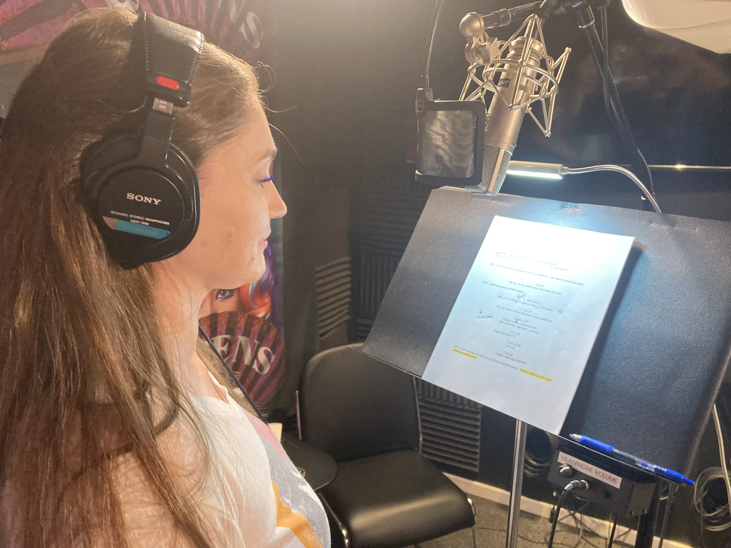 A woman with long reddish hair wearing black Sony headphones, sitting in a recording studio, focused on reading song lyrics on a music stand, with a large microphone in front of her.