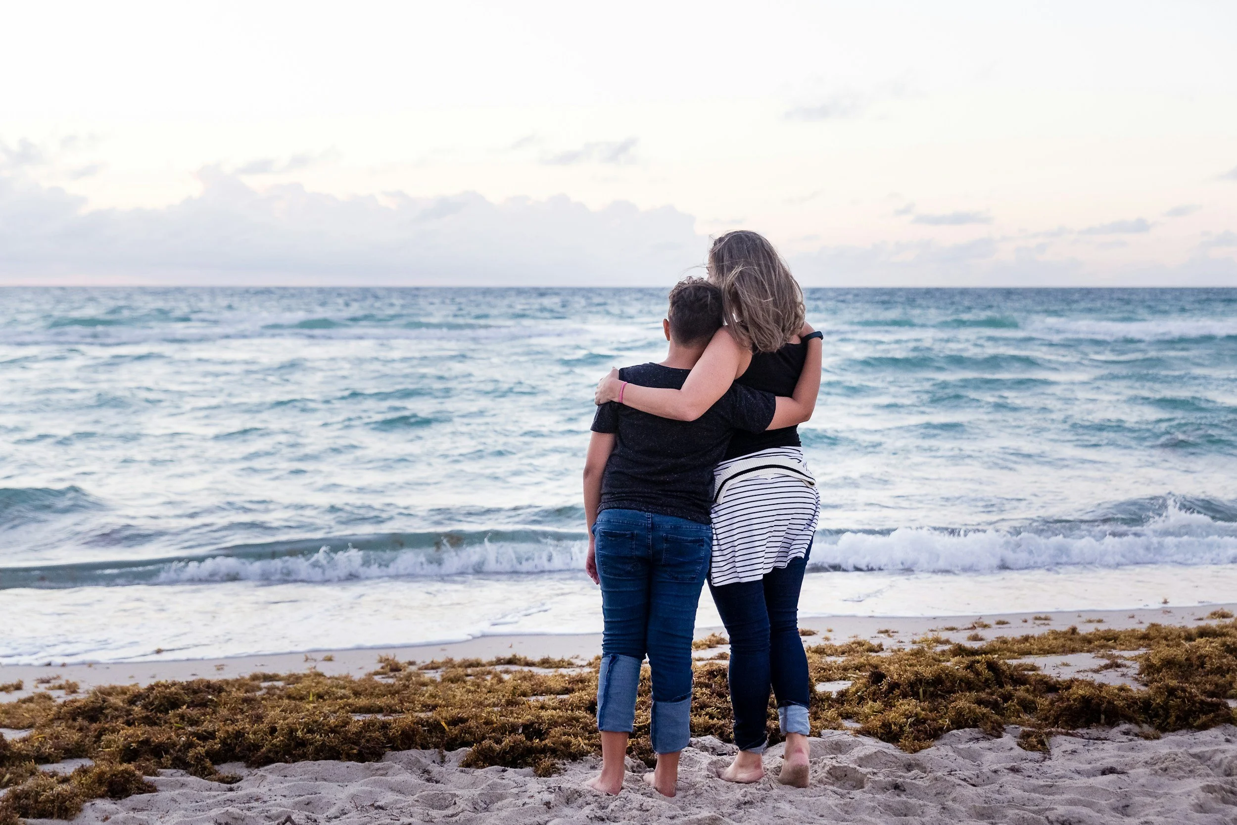 A woman and teen stand on the beach, hugging each other and looking at the ocean, with cloudy sky.
