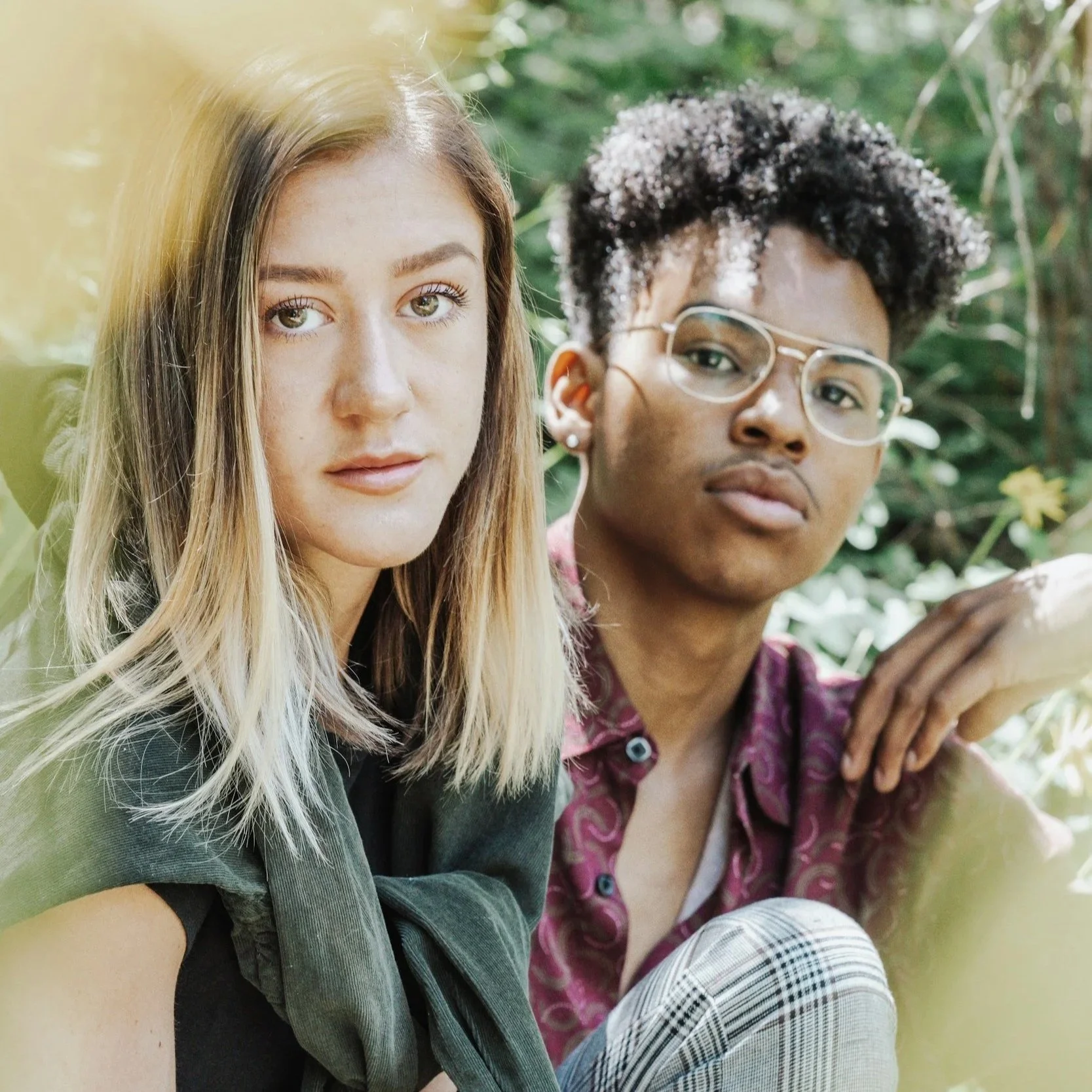 Two teenagers sitting outdoors in a natural setting, one with blonde hair and the other with curly hair and glasses, looking at the camera.