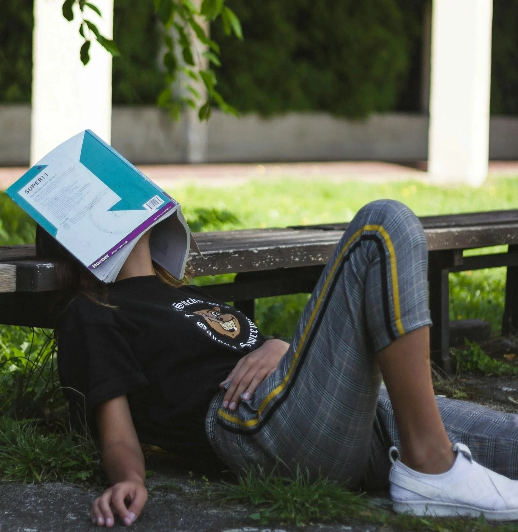 Person reclining on a park bench with a magazine covering their face, wearing a black t-shirt, plaid shorts, white sneakers, and a watch, with greenery in the background.