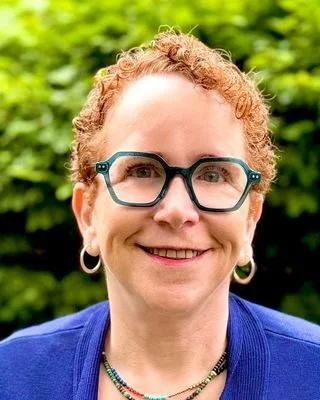 A woman with curly red hair, glasses, and gold hoop earrings standing outdoors with green foliage in the background.