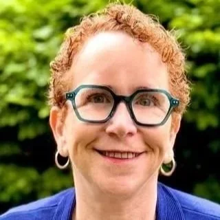 Carol Sampson, a woman with curly red hair, glasses, and hoop earrings smiling outdoors with green foliage in the background.