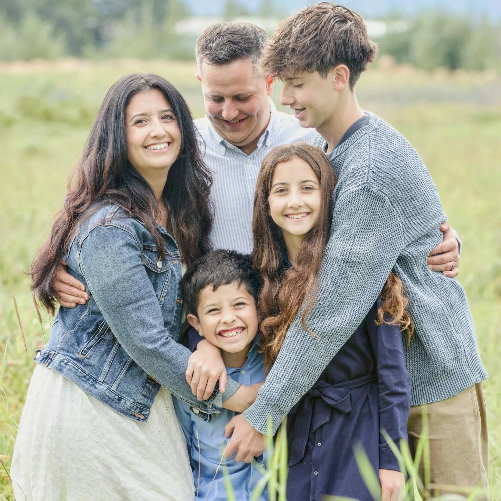 A happy family of five embracing outdoors in a green field, smiling and enjoying each other's company.